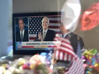 A man in the lobby of Senator John McCain's former office building watches a Fox News report as flowers and balloons from a makeshift memorial located outside the building are reflected in the building's glass window. (Robyn Beck / AFP)