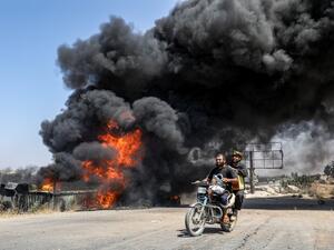 Men ride a motorcycle past a plume of smoke from a blaze at a vehicle gathering point for civilians fleeing from the south of Idlib province. (AFP/ File Photo)