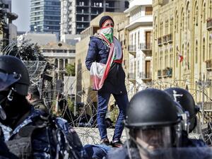An anti-government protester wearing a Lebanese national flag as a face bandana stands atop a concrete block behind riot police and ahead of a security barrier during a demonstration in the centre of the capital Beirut on January 27, 2020. JOSEPH EID / AFP