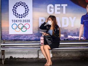 A woman wearing a facemask, amid concerns over the spread of the COVID-19 coronavirus, sits at a bus stop in front of a Tokyo 2020 Olympics advertisement in Bangkok on March 16, 2020. Mladen ANTONOV / AFP