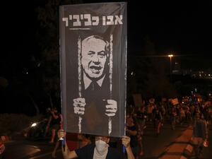 A protester wearing a face covering holds up a sign during a demonstration against the Israeli prime minister outside his official residence in Jerusalem, early on July 22, 2020. Israeli Prime Minister Benjamin Netanyahu, 70, was indicted in January for bribery, fraud and breach of trust in three cases. He denies all the charges and accuses the media and legal officials of a witch hunt. AHMAD GHARABLI / AFP