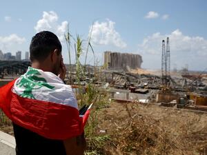 A man draped in a Lebanese flag reacts as he stands before the ravaged port of Lebanon's capital Beirut on August 9, 2020, in the aftermath of a colossal explosion that occurred days prior due to a huge pile of ammonium nitrate that had languished for years at a port warehouse. The huge chemical explosion that hit Beirut's port, devastating large parts of the Lebanese capital and claiming over 150 lives, left a 43-metre (141 foot) deep crater, a security official said. The blast Tuesday, which was felt acro