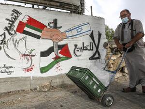A mask-clad man (COVID-19 coronavirus pandemic precaution) pushes a cart past a graffiti depicting hands with sleeves coloured in the UAE and Bahrain flags shaking hands with another with a sleeve bearing the Israeli flag, with text in Arabic reading (R to L) "no to normalisation" and "normalisation is betrayal", along a street in the flashpoint city of Hebron in the occupied West Bank on October 4, 2020. HAZEM BADER / AFP