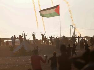 Palestinan protesters wave as they stand next to a large Palestinian flag with tear gas canisters falling above them, during a demonstration near the border with Israel east of Jabalia in the central Gaza Strip on May 15, 2018. MOHAMMED ABED / AFP