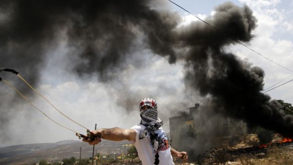Smoke billows from burning tires as a Palestinian protester uses a slingshot to hurl stones at Israeli troops, during clashes following a weekly protest against the expropriation of Palestinian land by Israel, in the village of Kfar Qaddum near the Jewish settlement of Qadumim (Kedumim), in the Israeli-occupied West Bank on July 19, 2019.  JAAFAR ASHTIYEH / AFP