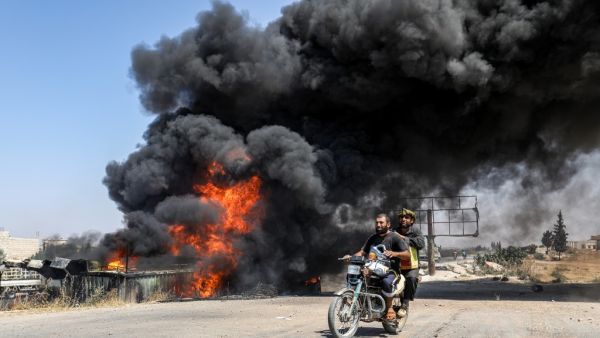 Men ride a motorcycle past a plume of smoke from a blaze at a vehicle gathering point for civilians fleeing from the south of Idlib province. (AFP/ File Photo)