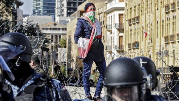 An anti-government protester wearing a Lebanese national flag as a face bandana stands atop a concrete block behind riot police and ahead of a security barrier during a demonstration in the centre of the capital Beirut on January 27, 2020. JOSEPH EID / AFP