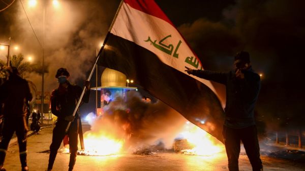 Masked anti-government protester wave the national flag before flaming tyres at a make-shift roadblock in the central Iraqi holy shrine city of Najaf on February 1, 2020. Iraq's president named former communications minister Mohammad Allawi as the country's new prime minister after an 11th-hour consensus among political blocs, but the streets seemed divided on his nomination. Haidar HAMDANI / AFP