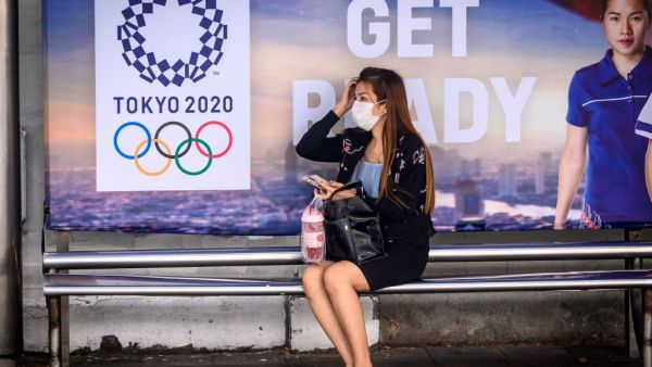 A woman wearing a facemask, amid concerns over the spread of the COVID-19 coronavirus, sits at a bus stop in front of a Tokyo 2020 Olympics advertisement in Bangkok on March 16, 2020. Mladen ANTONOV / AFP