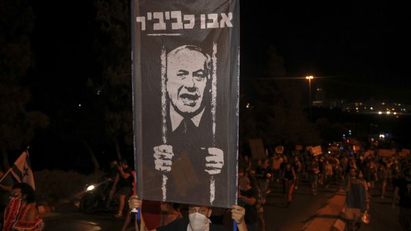 A protester wearing a face covering holds up a sign during a demonstration against the Israeli prime minister outside his official residence in Jerusalem, early on July 22, 2020. Israeli Prime Minister Benjamin Netanyahu, 70, was indicted in January for bribery, fraud and breach of trust in three cases. He denies all the charges and accuses the media and legal officials of a witch hunt. AHMAD GHARABLI / AFP