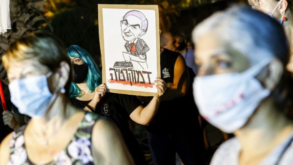 A protester, mask-clad due to the COVID-19 coronavirus pandemic, stands holding a sign showing a cartoon depicting Israeli Prime Minister Benjamin Netanyahu stepping on text dripping with red drops reading in Hebrew "democracy", during a demonstration against the Israeli government in the Mediterranean coastal city of Tel Aviv on August 1, 2020. JACK GUEZ / AFP