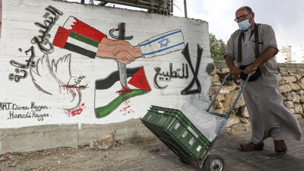 A mask-clad man (COVID-19 coronavirus pandemic precaution) pushes a cart past a graffiti depicting hands with sleeves coloured in the UAE and Bahrain flags shaking hands with another with a sleeve bearing the Israeli flag, with text in Arabic reading (R to L) "no to normalisation" and "normalisation is betrayal", along a street in the flashpoint city of Hebron in the occupied West Bank on October 4, 2020. HAZEM BADER / AFP