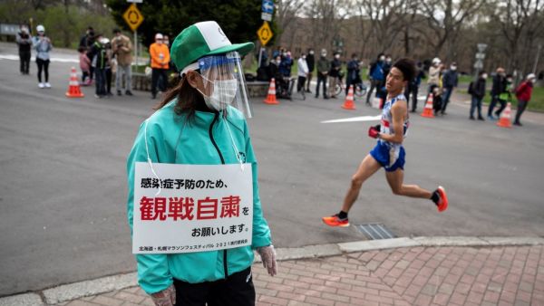 This file photo taken on May 5, 2021 shows a volunteer (L) holding a placard asking people to refrain from watching the competition to prevent the spread of the Covid-19 coronavirus while an athlete (R) competes in the half-marathon race which doubles as a test event for the 2020 Tokyo Olympics, in Sapporo. The public will be asked not to line the route of the Olympic marathon over fears that crowds of fans could spread coronavirus infections, Tokyo 2020 organisers said 