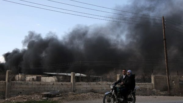 Syrians ride their motorbike as smoke billows in the background following reported air strikes on the rebel-held village of al-Chifouniya, on the outskirts of the capital Damascus, on March 4, 2016. (AFP/Amer Almohibany)