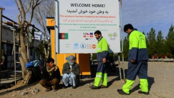 صورة تظهر المهاجرين الأفغان العائدين من إيران لدى جلوسهم تحت لافتة لمنظمة الهجرة الدولية في هرات

