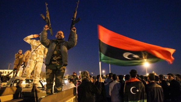 A Libyan man points two Kalashnikov assault rifles in the air as a national flag waves behind him during a celebration marking the sixth anniversary of the Libyan revolution. (AFP/File)