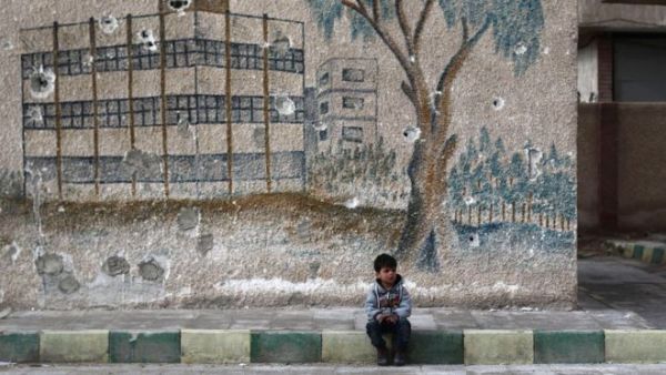 A Syrian child sits in front of a mural covered in bullet holes on the wall. (AFP/File)