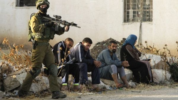 Israeli soldier keeps watch as Palestinians sit after the army entered Yatta earlier this week. (AFP/File)