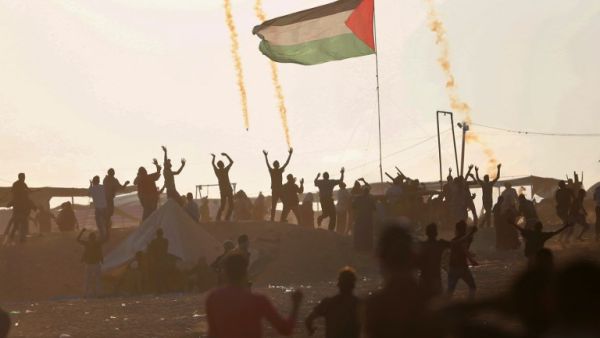 Palestinan protesters wave as they stand next to a large Palestinian flag with tear gas canisters falling above them, during a demonstration near the border with Israel east of Jabalia in the central Gaza Strip on May 15, 2018. MOHAMMED ABED / AFP
