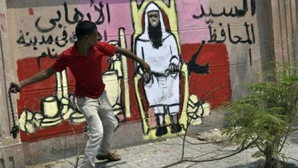 متظاهر يمسك بسلسلة حديدية أمام رسم ساخر على الجدار ضد المحافظ الجديد للاقصر
