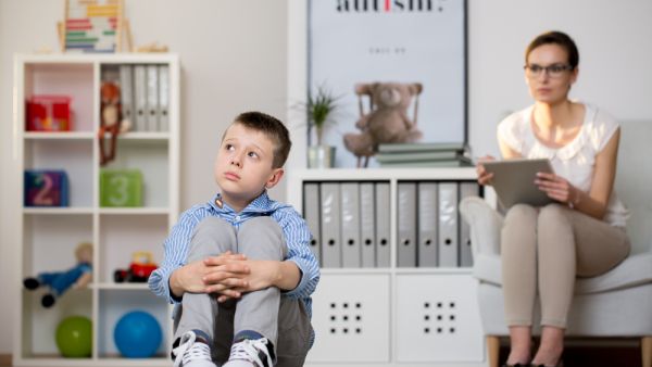 Psychologist in glasses is looking at kid sick of autism sitting on carpet in classroom. (Shutterstock/ File Photo)