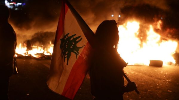 An Lebanese anti-government protester carries a Lebanese flag in front of burning tires blocking the main highway linking the city of Tripoli to Beirut at the coastal city of Byblos, north of the capital on November 13, 2019.  (AFP/ File Photo)