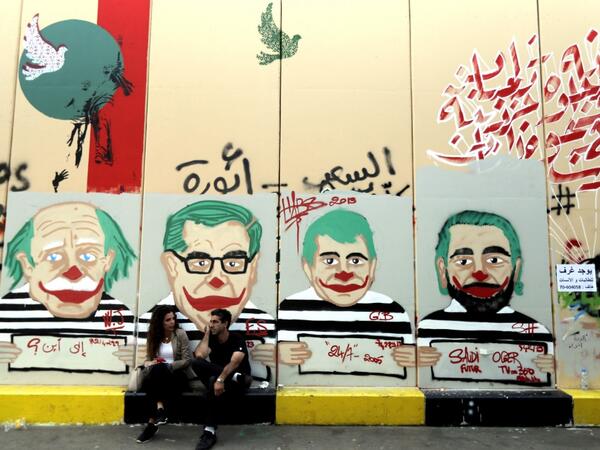 Anti-government protesters sit under graffiti depicting Lebanese politicians with joker faces sprayed on a wall in downtown Beirut on November 12, 2019. Banks and schools in Lebanon were closed today as protesters tried to prevent employees from clocking in at state institutions nearly one month into an anti-graft street movement. Unprecedented protests erupted across Lebanon on October 17, demanding the ouster of a generation of politicians seen by demonstrators as inefficient and corrupt.  JOSEPH EID / AF