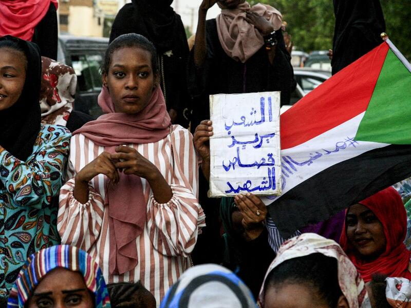 A Sudanese woman holds up a sign reading in Arabic "the people want retribution for the martyrs" next to a waving national flag defaced with the Arabic text "#CiviliaNow". (AFP/ File Photo)