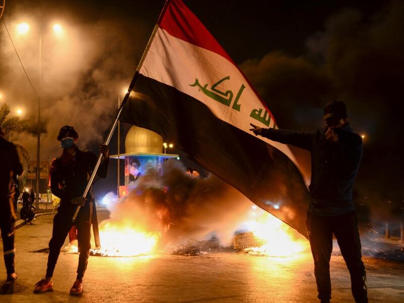 Masked anti-government protester wave the national flag before flaming tyres at a make-shift roadblock in the central Iraqi holy shrine city of Najaf on February 1, 2020. Iraq's president named former communications minister Mohammad Allawi as the country's new prime minister after an 11th-hour consensus among political blocs, but the streets seemed divided on his nomination. Haidar HAMDANI / AFP