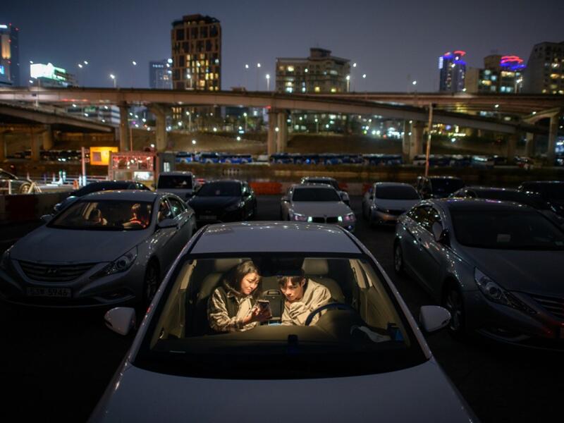 In a photo taken on March 21, 2020 a couple look at a mobile phone as they sit in a car at a screening at a drive-through cinema in Seoul. Box office numbers in South Korea -- which has 8,897 confirmed virus cases -- have plummeted in recent weeks due to the epidemic, with authorities urging the public to avoid large crowds. But at drive-in cinemas, moviegoers can enjoy a movie from the comfort of their cars, parked in front of a large outdoor screen.  Ed JONES / AFP