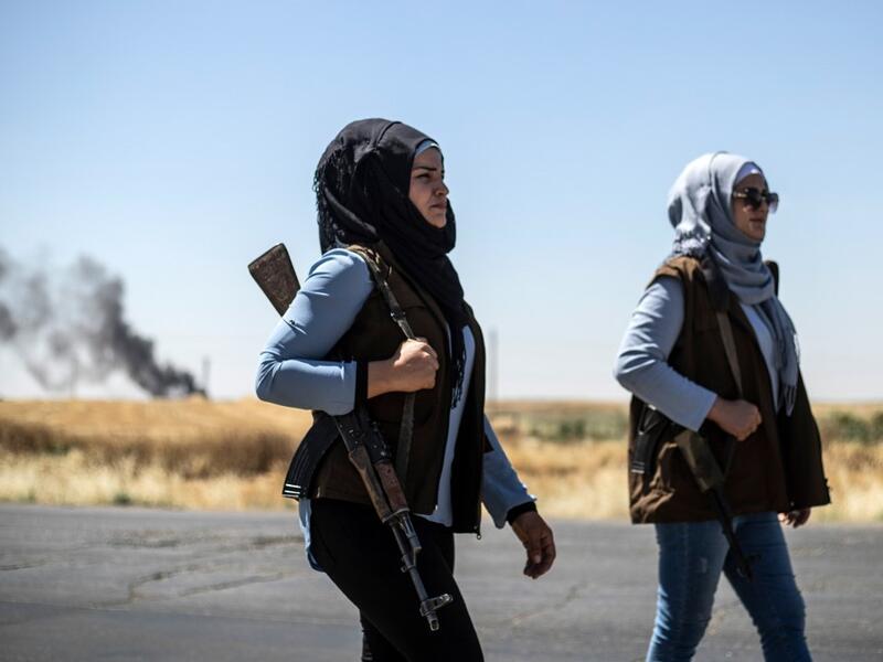 Smoke billows from an oil field as Kurdish female volunteers, from the newly formed Community Protection Forces, guard a wheat field, against threats by jihadists to burn the crops, during harvest season on June 13, 2020, in the countryside east of Qamishli in Syria's northeastern Hasakah province. Delil SOULEIMAN / AFP