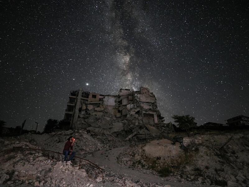 This long-exposure picture taken early on June 27, 2020 shows a man smoking past buildings destroyed by prior bombardment in the town of Ariha in Syria's rebel-held northwestern Idlib province, as the Milky Way galaxy is seen in the night sky above. Omar HAJ KADOUR / AFP