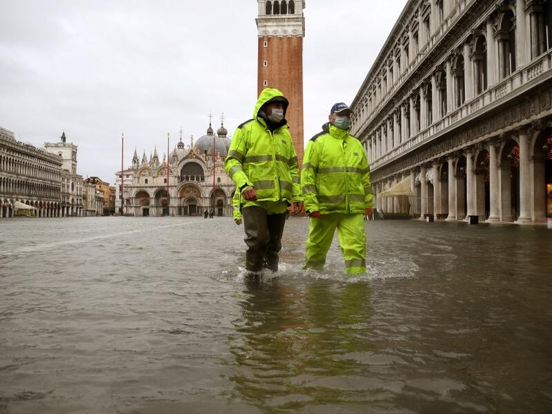 Men wearing waterproof gear walk across a flooded St. Mark's Square on December 8, 2020 following a high tide "Alta Acqua" event following heavy rains and strong winds, and the mobile gates of the MOSE Experimental Electromechanical Module that protects the city of Venice from floods, were not lifted. ANDREA PATTARO / AFP
