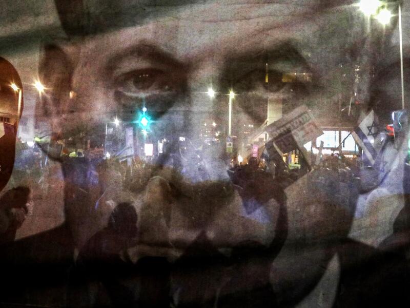 In this file photo taken on August 11, 2018, silhouettes of demonstrator are seen through a giant poster depicting Israeli Prime Minister Benjamin Netanyahu as Arab Israelis and their supporters march in a rally to protest against the 'Jewish Nation-State Law' in the Israeli coastal city of Tel Aviv. Israel's Prime Minister Benjamin Netanyahu returns to court on February 8, 2021 to formally respond to the corruption charges filed against him, as his trial enters an intensified phase six weeks before he face