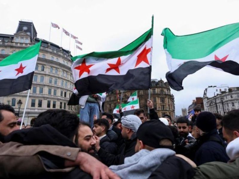 متظاهرون يرفعون أعلام الثورة السورية في ساحة عامة وسط تجمع حاشد، مع مبانٍ تاريخية في الخلفية.