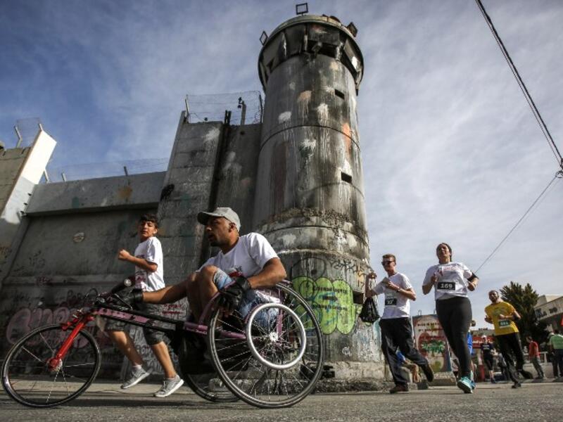 A participant rides in a wheelchair with others running along Israel's controversial separation barrier, which divides the West Bank from Jerusalem, in the biblical town of Bethlehem during the 6th International Palestine Marathon on Mar. 23, 2018. 
(Musa AL SHAER / AFP)
