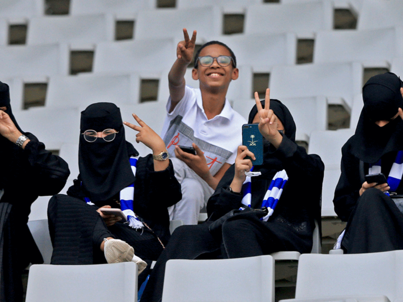مشجعو كرة القدم السعوديون ، بمن فيهم النساء ، يبثون لفتة النصر وهم ينتظرون فريقهم في المدرجات قبيل مباراة كرة القدم الفاصلة في دوري أبطال آسيا بين الأهلي السعودي والهلال على ملعب جامعة الملك سعود بالعاصمة السعودية الرياض في 13 أغسطس 2019.