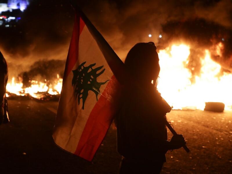 An Lebanese anti-government protester carries a Lebanese flag in front of burning tires blocking the main highway linking the city of Tripoli to Beirut at the coastal city of Byblos, north of the capital on November 13, 2019.  (AFP/ File Photo)