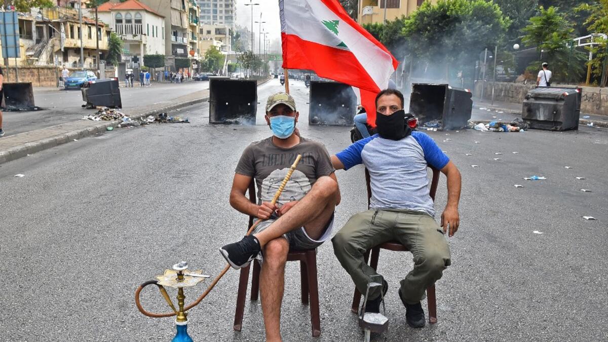 A masked man smokes a waterpipe (also known as shisha or hookah) while another holds up a Lebanese national flag as they sit together before overturned dumpsters blocking a road in the centre of the Lebanese capital Beirut on October 18, 2019 amidst ongoing protests against dire economic conditions. Public anger has simmered since parliament passed an austerity budget in July to help trim a ballooning deficit and flared on October 17 over new plans to tax calls on messaging applications such as WhatsApp, fo
